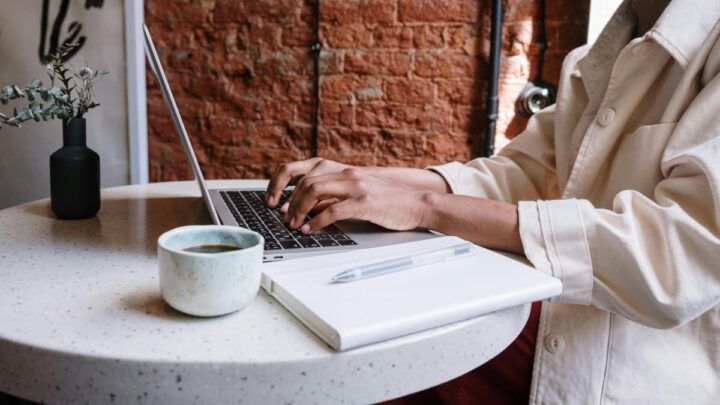 Uma pessoa a tirar notas: veem-se umas mãos em posição de digitação sobre um teclado de um computador portátil. Ao lado, um café e uma bloco de notas físico. 